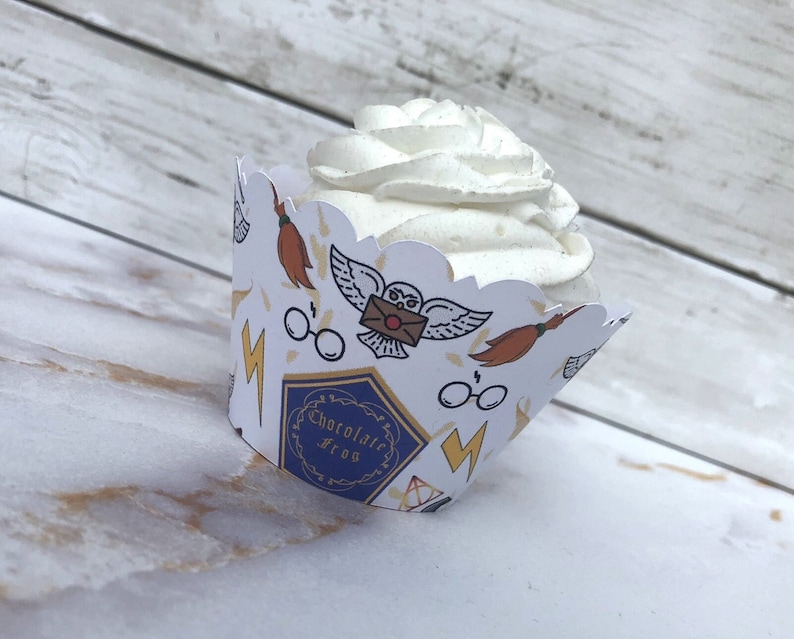 May include: A cupcake in a decorative paper wrapper. The white wrapper has a scalloped edge and is printed with images of a broom, owl, glasses, and lightning bolt. The wrapper also has the text "Chocolate Frog" in a blue and gold design.