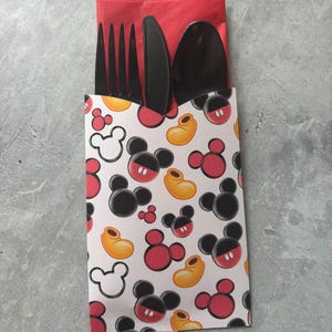 May include: A black fork, knife, and spoon set inside a red napkin. The napkin and utensils are in a white paper sleeve with a pattern of Mickey Mouse heads, red shorts, and yellow shoes.