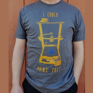 May include: A gray t-shirt with a yellow and blue illustration of a 3D printer. The text "I could print that" is printed in yellow on the shirt.