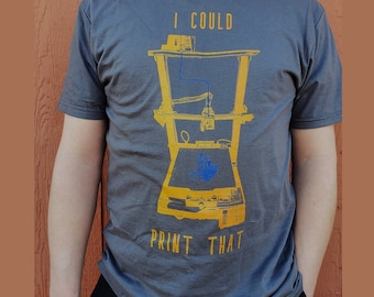 Puede incluir: Una camiseta gris con una ilustración amarilla y azul de una impresora 3D. El texto "I could print that" está impreso en amarillo en la camiseta.