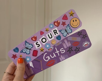 Könnte beinhalten: Zwei lila Lesezeichen mit bunten Aufklebern und den Worten "Sour Guts" darauf.