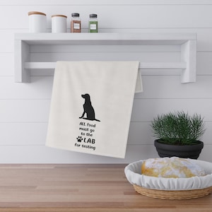 May include: A white kitchen towel with a black silhouette of a dog and the text "ALL food must go to the LAB for testing." The towel is hanging on a white shelf with other kitchen items.
