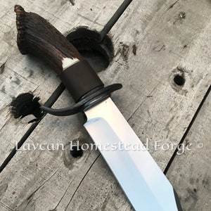 May include: A large, fixed blade knife with a brown antler handle and a silver blade. The blade is sharp and has a slight curve at the tip. The knife is resting on a wooden surface. The text "Laycan Homestead Forge" is visible on the surface.