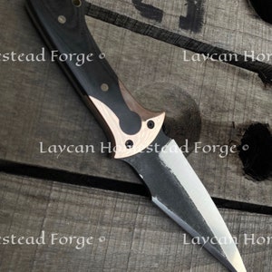 Op de afbeelding: Een mes met een zwart en koperen handvat en een scherp, puntig blad. Het mes is gemaakt door Laycan Homestead Forge.
