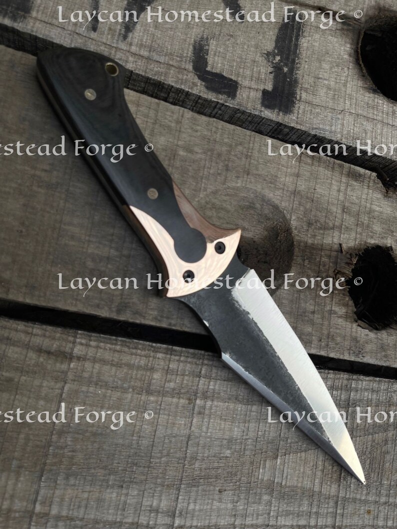 Op de afbeelding: Een mes met een zwart en koperen handvat en een scherp, puntig blad. Het mes is gemaakt door Laycan Homestead Forge.