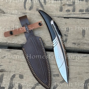 Puede incluir: Una daga artesanal con una hoja plateada pulida y un mango negro, junto a una funda de cuero marrón. La funda tiene costuras decorativas y una trabilla para el cinturón. El texto "Lycan Homestead Forge" es visible.