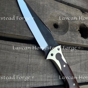 Op de afbeelding: Een zilveren en zwart mes met een bruin houten handvat. Het handvat heeft een messing inlay en een zilveren klinknagel. Het mes ligt op een houten oppervlak.