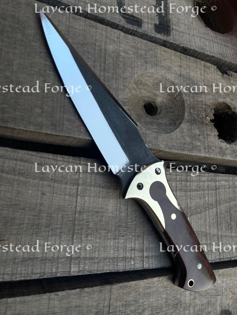 Op de afbeelding: Een zilveren en zwart mes met een bruin houten handvat. Het handvat heeft een messing inlay en een zilveren klinknagel. Het mes ligt op een houten oppervlak.