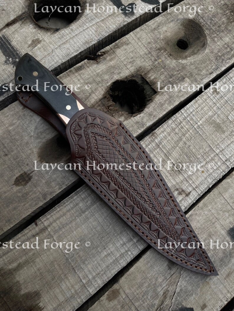 Op de afbeelding: Een bruine leren schede met ingewikkelde gegraveerde ontwerpen en stiksels. De schede is voor een mes met een zwart handvat en een zilveren lemmet. De tekst "Laycan Homestead Forge" is zichtbaar op de schede.