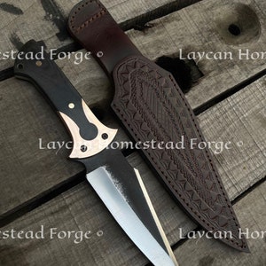 Op de afbeelding: Een zwart en bruin mes met een zilveren lemmet en een leren schede. De schede heeft een decoratief patroon en de tekst "Laycan Homestead Forge" is erop gegraveerd.