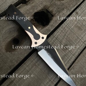 Op de afbeelding: Een mes met een zwart en bronzen handvat en een scherp, puntig blad. Het mes ligt op een houten oppervlak. De tekst "Laycan Homestead Forge" is zichtbaar op het blad.