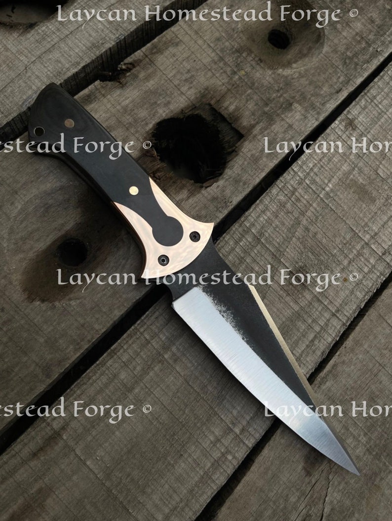 Op de afbeelding: Een mes met een zwart en bronzen handvat en een scherp, puntig blad. Het mes ligt op een houten oppervlak. De tekst "Laycan Homestead Forge" is zichtbaar op het blad.
