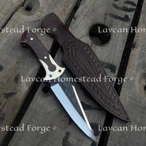 Op de afbeelding: Een bruine leren schede met een decoratief patroon en een zilveren mes met een bruin houten handvat. Het mes en de schede liggen op een houten oppervlak met de tekst "Laycan Homestead Forge" erop gedrukt.