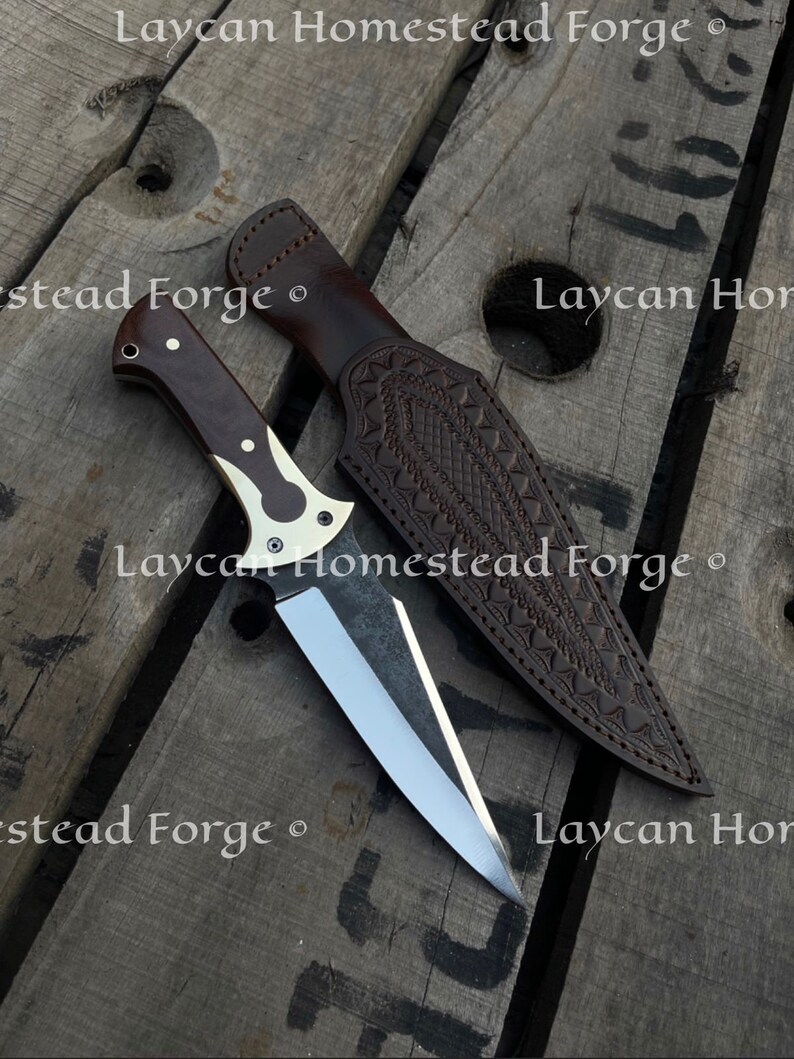 Op de afbeelding: Een bruine leren schede met een decoratief patroon en een zilveren mes met een bruin houten handvat. Het mes en de schede liggen op een houten oppervlak met de tekst "Laycan Homestead Forge" erop gedrukt.