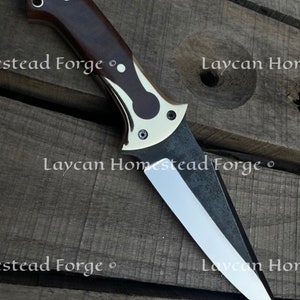 Op de afbeelding: Een close-up van een mes met een bruin houten handvat en een zilveren metalen beschermkap. Het blad is zilver en heeft een zwarte afwerking. Het mes ligt op een houten oppervlak. De tekst "Laycan Homestead Forge" is zichtbaar op het blad.