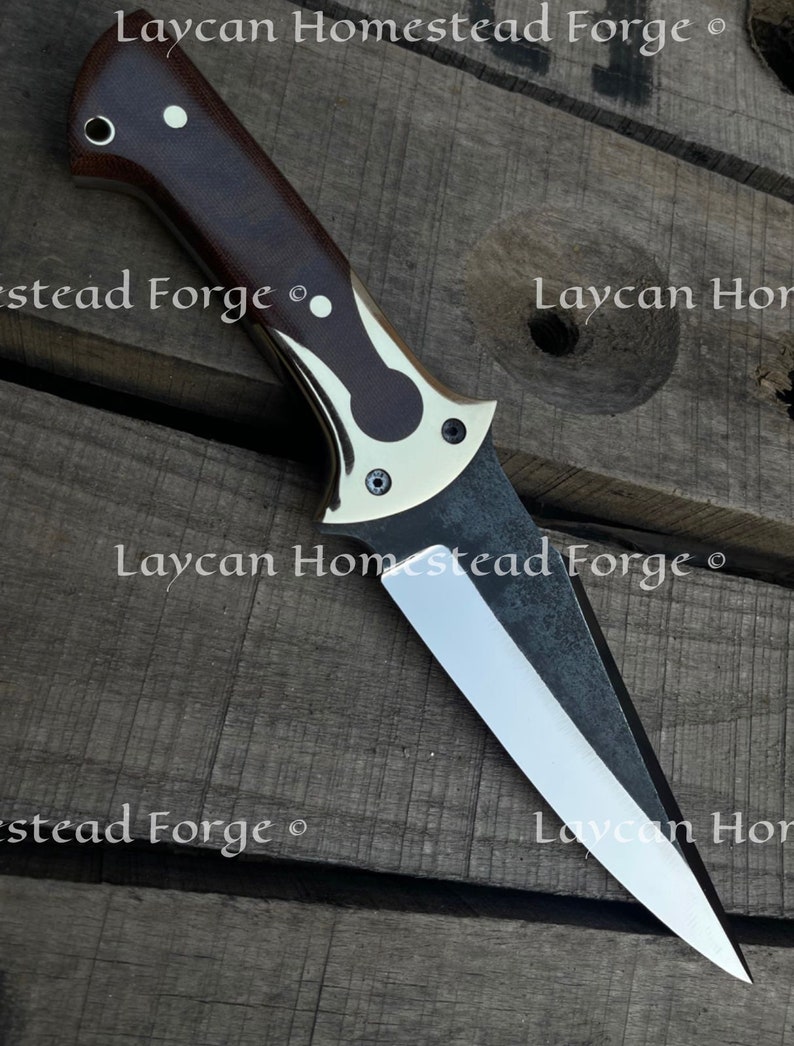 Op de afbeelding: Een close-up van een mes met een bruin houten handvat en een zilveren metalen beschermkap. Het blad is zilver en heeft een zwarte afwerking. Het mes ligt op een houten oppervlak. De tekst "Laycan Homestead Forge" is zichtbaar op het blad.