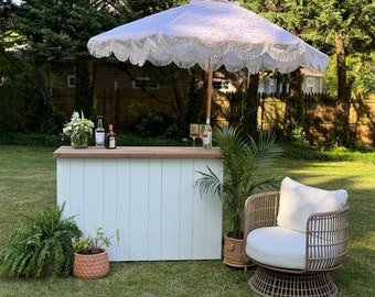 Può includere: Banco bar in legno bianco con un piano in legno, due bottiglie e due bicchieri. Una sedia in vimini bianca con un cuscino bianco si trova di fronte al bancone. Una palma e una felce sono in vasi su entrambi i lati del bancone. Un ombrello bianco e beige con frange è sopra il bancone.