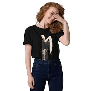 以下が含まれることがあります： 魚を持っている人のグラフィックプリントが施された黒いTシャツ。その人は黒いシャツとズボンを着用しています。