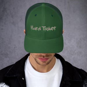 Könnte beinhalten: Eine grüne Trucker-Kappe mit einem pinkfarbenen "Hard Ticket"-Stickerei auf der Vorderseite.