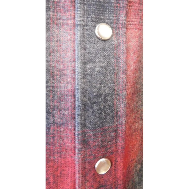 May include: A long-sleeved shirt featuring a red and charcoal gray plaid pattern. The garment includes a vertical button placket down the center with two visible round, metallic-toned snap buttons that have a smooth, reflective surface finish.