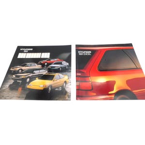 May include: Two brochures featuring Hyundai cars. The left brochure has the text "HYUNDAI 1991" and showcases several car models in various colors. The right brochure displays a red Hyundai Excel with the text "HYUNDAI 1991 EXCEL".