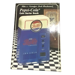 May include: A Pepsi-Cola Coin Sorter Bank in a box. The blue and white bank features the Pepsi-Cola logo, the text "Drink Pepsi-Cola," "Ice Cold," and "Take a Nostalgic Look Backward." The box displays a checkerboard pattern and a framed picture.