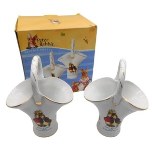 May include: Two white ceramic baskets with handles, featuring gold trim and illustrations of Peter Rabbit and Benjamin Bunny. The baskets are from the Beatrix Potter collection and are displayed with their original box.