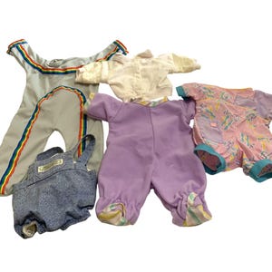 May include: A collection of doll clothing including a light gray jumpsuit with rainbow stripes, a white long-sleeve top, a blue denim-style overall with a small rectangular label, a solid lavender footed sleeper, and a pink patterned short-sleeve romper.