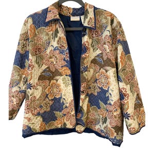 May include: A quilted jacket featuring a multi-colored floral and paisley pattern in shades of navy blue, tan, brown, and cream. The garment has a collared neckline, long sleeves, and a front button closure with a solid navy blue interior lining.