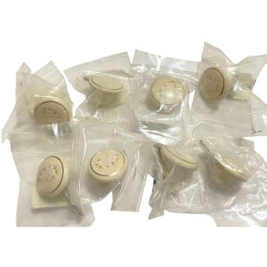 May include: A collection of eight round, off-white cabinet knobs, each individually packaged in a clear plastic bag. The knobs feature a raised, embossed floral pattern on the circular face and include a metal screw attachment on the back.