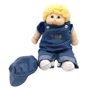 May include: A vintage doll with a soft, plush body, curly blonde hair, and blue eyes. The doll wears a denim blue overall with the word "Cuddlie" embroidered on the pocket, a blue shirt, and a matching denim cap.