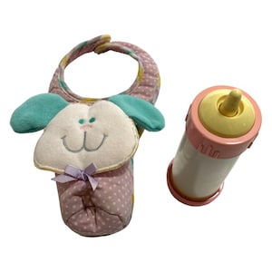 May include: A pink and white polka-dot fabric bib featuring a plush animal face with teal ears and a small purple ribbon bow, paired with a plastic toy baby bottle that has a pale yellow nipple and a pink cap and base with a white cylinder.