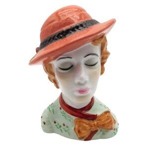 May include: A ceramic figurine of a woman's head and shoulders. She wears a coral-colored hat with a black band, a red scarf, and a green top with a bow. The woman's eyes are closed, and her lips are painted pink.