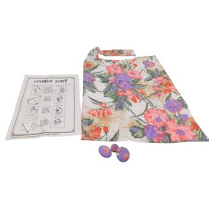 May include: A white scarf featuring a vibrant floral pattern in shades of orange, purple, and green, accompanied by three matching fabric-covered buttons and an instructional paper sheet titled creative scarf with diagrams for styling.