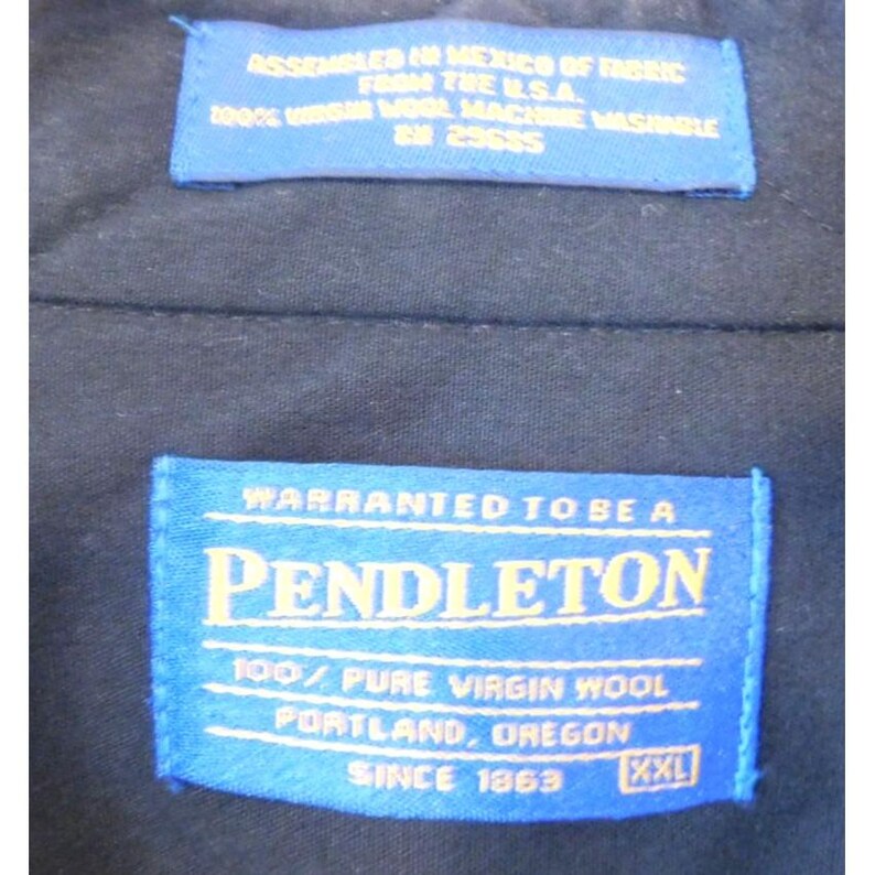 May include: A dark navy blue garment interior featuring two blue rectangular fabric labels with yellow text. The top label reads ASSEMBLED IN MEXICO OF FABRIC FROM THE U.S.A. 100% VIRGIN WOOL MACHINE WASHABLE RN 29685. The bottom label reads PENDLETON.