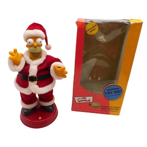 May include: A Homer Simpson figure dressed in a red and white Santa Claus suit with a matching hat, standing on a red circular base. Beside it is a rectangular cardboard box with a clear window, featuring the text The Simpsons and Try Me.