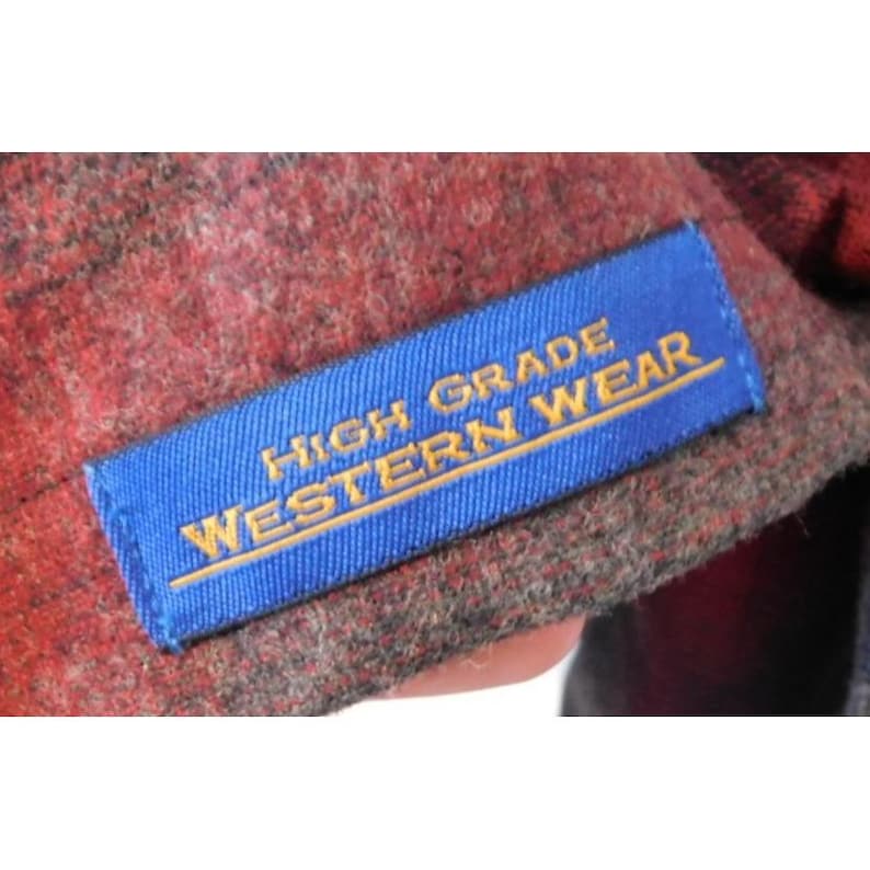 May include: A red and black plaid fabric garment featuring a rectangular blue label with yellow embroidered text that reads HIGH GRADE WESTERN WEAR. The fabric has a textured, woven appearance with visible fibers throughout the material.