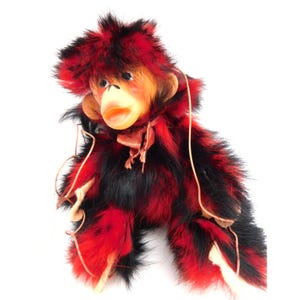May include: A plush monkey puppet featuring long, shaggy black and red faux fur. The monkey has a molded plastic face with a light tan complexion, a small open mouth, and brown eyes. Thin tan leather strings are attached to the limbs for movement.