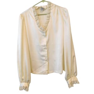 May include: A long-sleeved off-white blouse featuring a V-neckline with delicate lace trim, a front button closure with three small round buttons, and matching lace-trimmed cuffs on the sleeves. The garment has a smooth, satin-like finish.