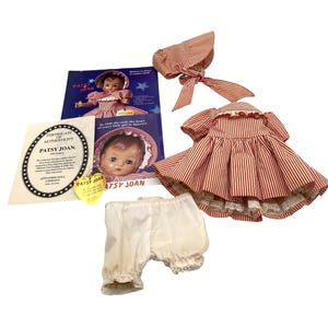 May include: A Patsy Joan doll set including a red and white striped dress with a ruffled collar, a matching bonnet with ribbon ties, and white bloomers with lace trim. Also included are a certificate of authenticity and promotional paper materials.