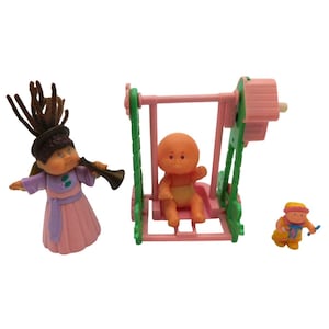 May include: A set of three small plastic figurines including a doll with brown yarn hair wearing a pink dress and holding a dark horn, a peach-colored baby doll sitting on a pink and green swing set, and a tiny baby doll in a yellow outfit.