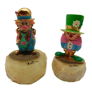 May include: Two clown figurines, each standing on a light-colored stone base. One clown wears a gold hat and blue overalls, the other a green top hat and blue jacket. Both have painted faces and are detailed with gold accents.