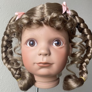 May include: A doll head with light brown hair styled in two braids and ringlets. The doll has brown eyes, rosy cheeks, and a neutral expression. Pink bows adorn the braids. The doll's hair is styled with a center part.