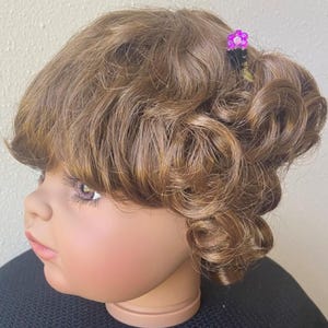 Puede incluir: Cabeza de muñeca con cabello castaño rizado y un pequeño accesorio para el cabello de flores moradas. La muñeca tiene tez clara, labios rosados y sombra de ojos rosa. La muñeca lleva un suéter negro.