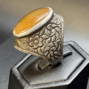 Puede incluir: Un anillo de plata con una piedra de ámbar ovalada engastada en el centro. La banda del anillo está finamente grabada con diseños florales.