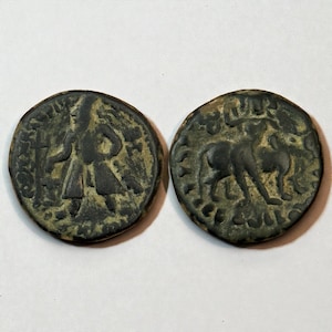 May include: Two antique bronze coins. One coin features a standing figure with outstretched arms and a long robe. The other coin shows a depiction of an animal, possibly a bull or a cow. Both coins have intricate details and are set against a white background.