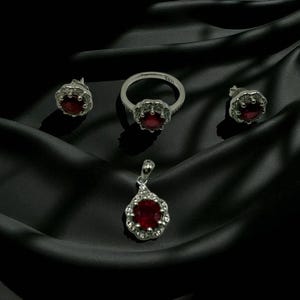 Könnte beinhalten: Ein Schmuckset mit Ring, Anhänger und Ohrringen. Jedes Teil hat eine silberne Oberfläche und einen zentralen roten Edelstein, der von kleineren, klaren Steinen umgeben ist. Das Set wird auf einem schwarzen, strukturierten Stoff präsentiert.