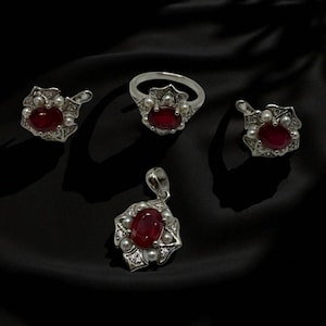 May include: A jewellery set comprising a ring, pendant, and earrings. Each item has a silver-coloured setting with a large, oval, deep red gemstone at the centre, encircled by small white pearls and clear accent stones. The set is displayed on black fabric.