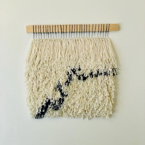 May include: A white and black woven wall hanging with a fringe border. The hanging is made of yarn and has a geometric design.