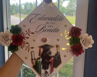 May include: A white graduation cap decorated with gold glitter, gold leaf, and red, green, and white paper flowers. The cap features a printed image of a woman in a graduation gown and cap, holding a diploma and a Mexican flag. The text "Educadita Me Veo Mas Bonita" is printed on the cap. The text "2 Out of 3" is printed on the bottom of the cap.